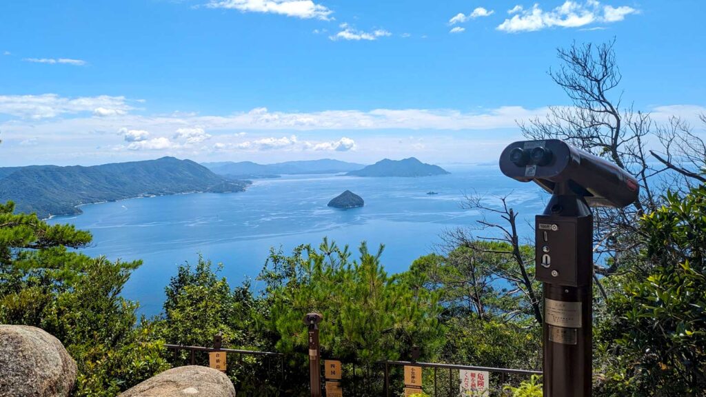 the view from the Shishiiwa Observation deck. It's a fenced in lookout that used the surroundings. 

Binoculars sit on a stand for visitors to look out at the view beyond. 

A view of sea and land, small and large mountainous islands