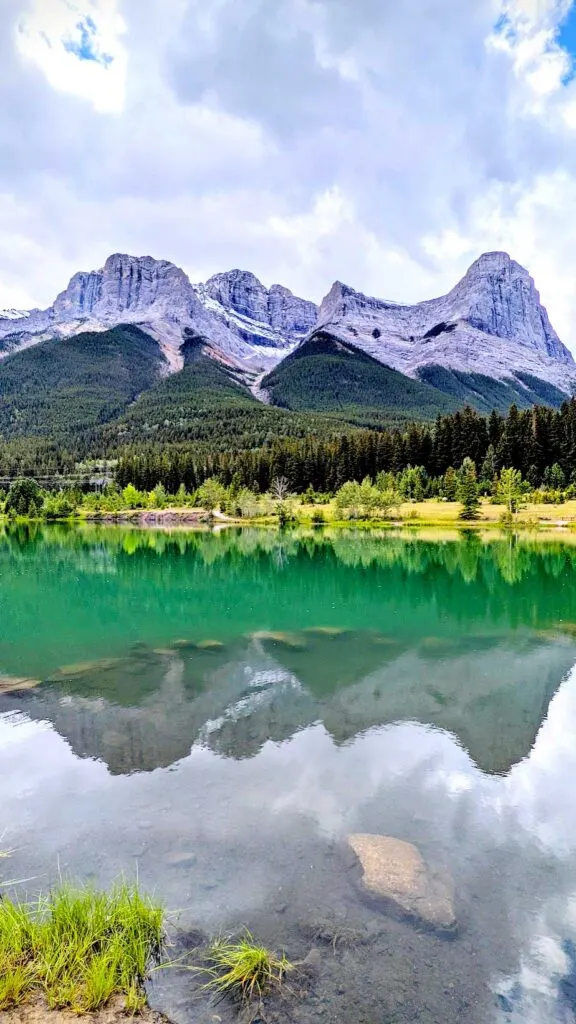 the incredible view from quarry lake park in canmore alberta. A vertail portrait that leads you from the grassy sands below, towards the extremely calm lake giving you a nearly perfect reflection in the clear teal waters before you stand in awe at the mountain range above. The clouds provide a rather ominous feel to the photograph, as they look like a storm could be brewing behind.