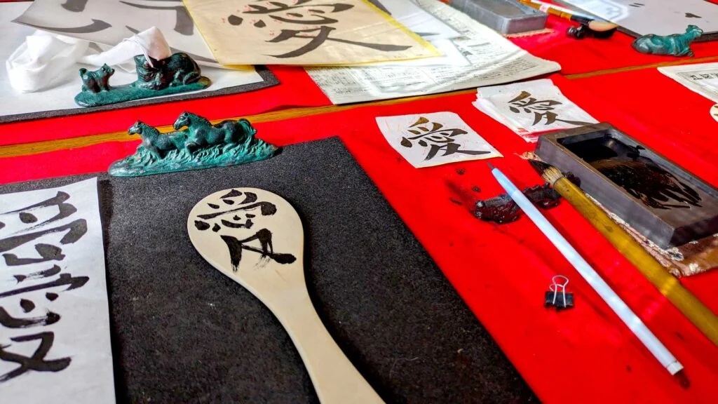 looking at a table with a bright red table cloth. There are a few unique items on the table, meant for traditional calligraphy. There's a small pot with black ink. Two paint brushes are on rests. There are a few small pages with kanji symbols. There's black paper on the edge, held down by a green dragon paperweight. On top sits a wooden rice scoop with kanji written on it