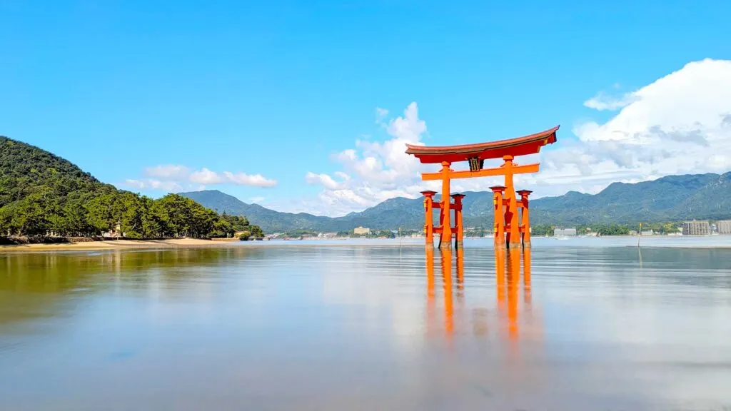 the magnificent torii gate in the middle of the water. A beautiful bright red gate standing tall in the water. The photo was taken with a long exposure so the water is perfectly smooth below, allowing for an almost perfect reflection, you can only see the pillars. Behind the gate is the Hiroshima prefecture mountains.