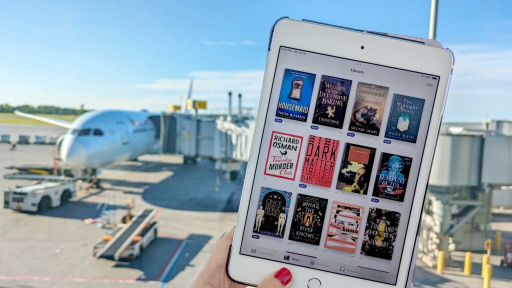 a woman with red nail polish holds up an ipad mini open on the ibook app filled with a digital book library of at least 12 titles on the screen. Behind the ipad is the plane shes about to board on a sunny evening