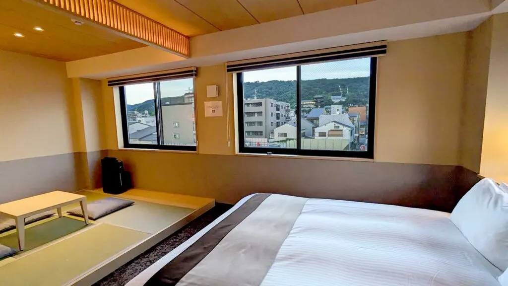hotel room in rinn gion yasaka kyoto. you see the large queen size bed eating up the right side of the image. In front of the bed is a platform with tatami floors and a small table. There are two large windows overlooking gion, you can even see the 5 story pagoda and the mountains surrounding kyoto