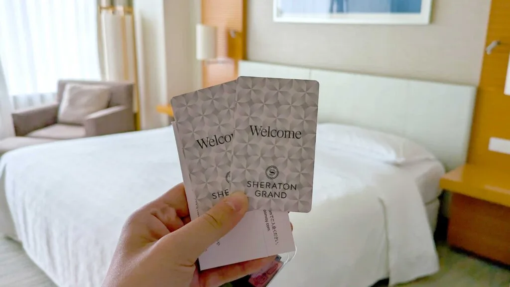 a hand in the middle of the frame holding up up two room keys. They're grey and white pattern with Shertaon Grand written on them. Behind the room keys is a bed, and a comfy looking armchair