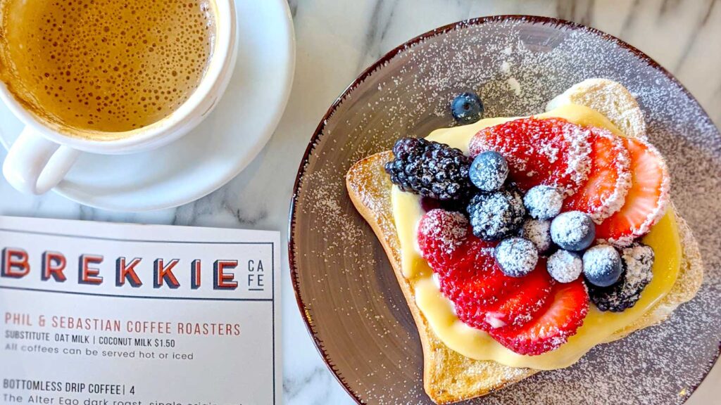 a photo take from above of a brekkie menu, written all in capital letters in red and black. There's a cappuccino in a white mug with a white saucer on top. Next to the menu on the right is a brown speckled plate with a piece of french toast topped with yellow custard, and the freshest berries, and everything has been sprinkled with powdered sugar to top it off! 