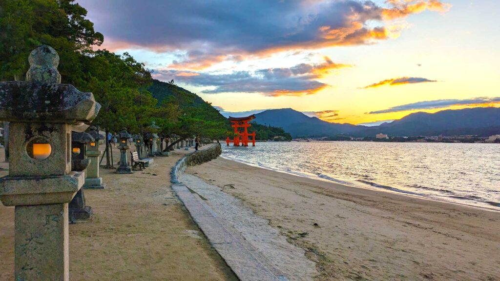 the view along the beach in miyajima island at sunset. Stone lanterns stand in a line, along the edge of the path and the start of the beach all the way down leading your way to the main point of interest: the otorii gate in the distance. The first stone lantern s actually lit up in the middle, with an orange light. It matches the orange of the torii gate all the way, standing in the middle of the water in the distance. There are mountains along the back edge of the image, continuing off into the right. Behind the mountains is the end of of sun set, orange and yellow colours burst from behind the mountain, changing to blue and when they meet the clouds in the sky change again to a pinkish hue. The colours are slightly reflected in the water, adding a bit of symmetry to the image