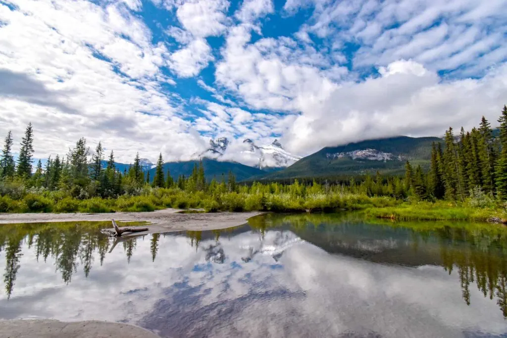 a wide angle view of the three sisters near canmore alberta. A calm lake leads your eye to the reflection of the three sisters, towering above it. It's a cloudy day, the blue sky is able to peak through behind the clouds, but the three peaks of the three sisters struggle. They do manage do break through enough to see that there are in fact three peaks next to each other. Trees line the base of the mountains. 