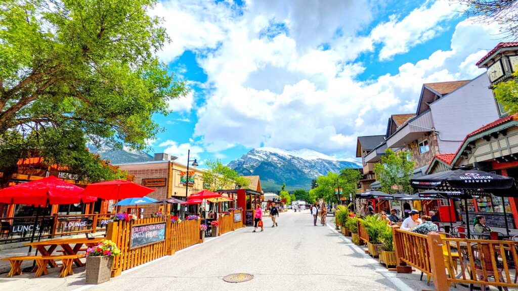 vibrant pedestrian only street in canmore alberta. The street is lined on both sides with fenced in terraces for people to enjoy lunch and dinner. The left side has bright red umbrellas open to provide shade, the large tree above them aren't enough. The right side only has small baby trees, and lack umbrellas opened up