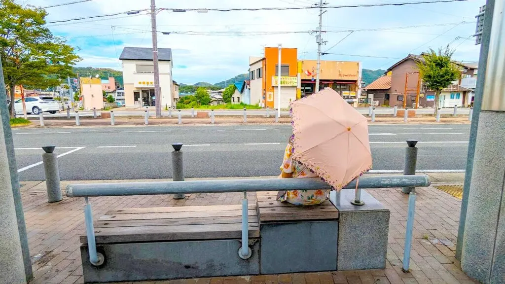 a woman sits on a concrete bench at a bus stop, her back is to the camera and she has a pink umbrella open to shield herself from the hot japan summer sun. This isn't a very popular area, there aren't any cars on the street and only a handful of older buildings across from the bus stop