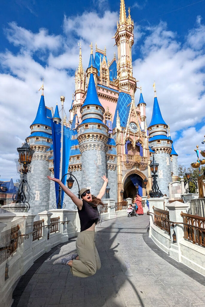 a woman jumping in mid air at the entrace of the castle in magic kingdom on the ramp going up to the blue and pink magical castle. Magically, there's no one behind her, just some blurs at the entrance but you can barely see them. 