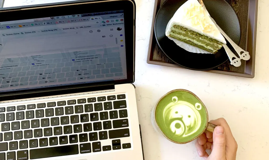 working at a cafe. A laptop is open, a hand comes into the frame to pick up the mug, it's filled with bright green matcha with latte art in the shape of a cute bear. There's also a matcha cake on the table to be enjoyed