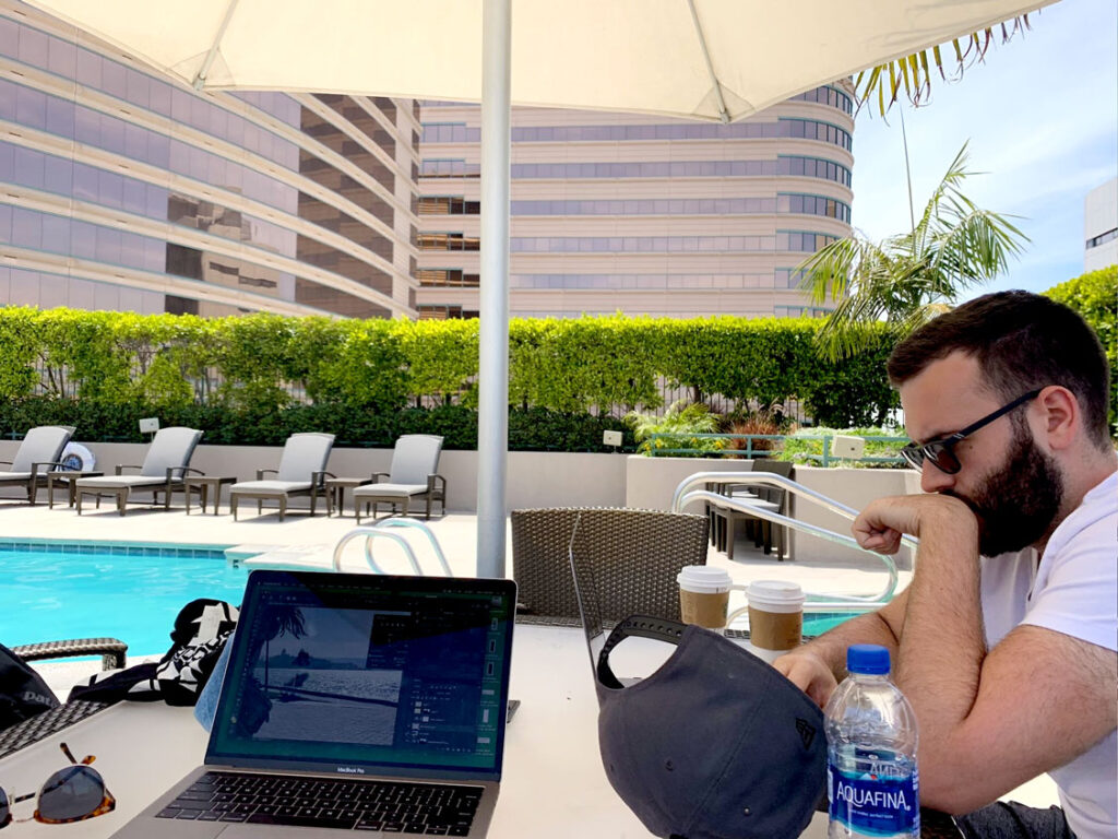 working hard at the pool/ A laptop is open on photoshop and a man is looking down concerned. They sit at an outdoor table, poolside, with lounge chairs and tall building behind on a bright and sunny day.