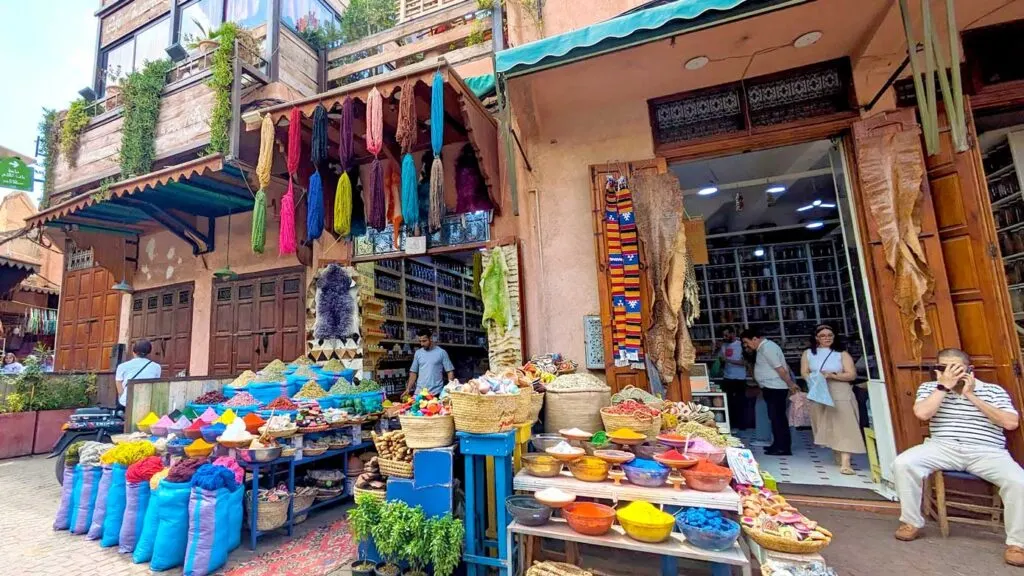 a vibrant shop at the marrekech market i morocco. The vendor has set out outside of the shop different spices, yarn, and very vibrant powders. The shopkeeper is sitting outside talking on a cellphone while tourist are inside his shop