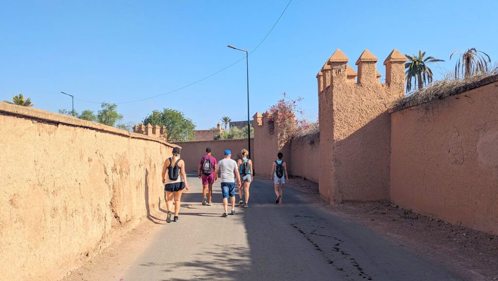 a snapshot of the active walking group from the club med marrakech. A group of people are walking ahead in a group, they're dressed in workout attire with shorts, t shirts and hats - on a mission to move! They are on a street thats  lined with an old red clay walls, throughout the walls on the right side there are medieval looking towers.