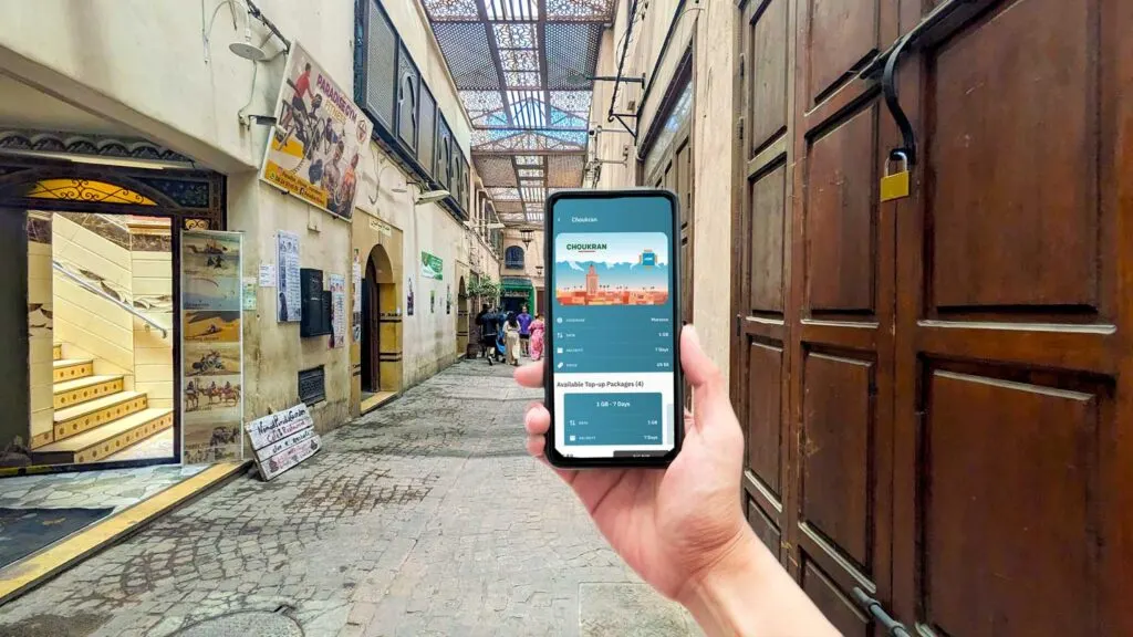 a desolate market street in Marrakech, ther eisn't a lot of colour, just a deep brown closed door on the right. In the front, a hand is holding up a phone with it open to Airalo App showing Choukran - the Moroccan data plan 