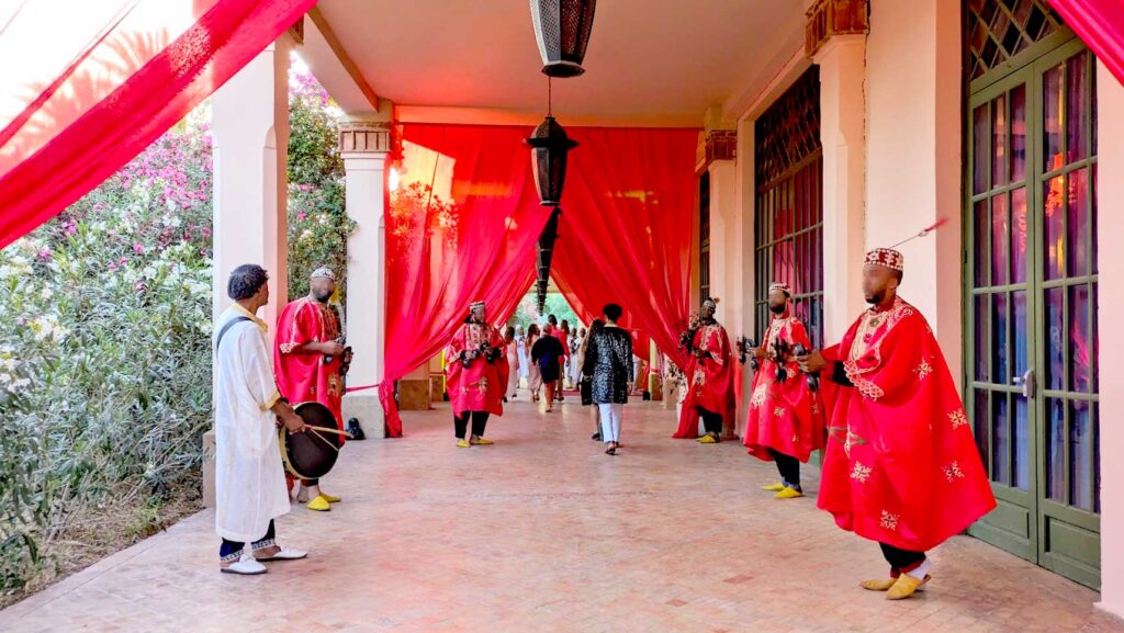 a party is happening, welcoming guests to the club med Marrakech. A line up of four staff stand on either of the open corridor, they're are dressed in traditional Moroccan attire, a red and gold embroidered overcoat, wearing a fez and playing an instrument. They're smiling and dancing with guests as they walk by. There are also sheer red drapes hanging from the ceilings, going almost all the way to the floor. 