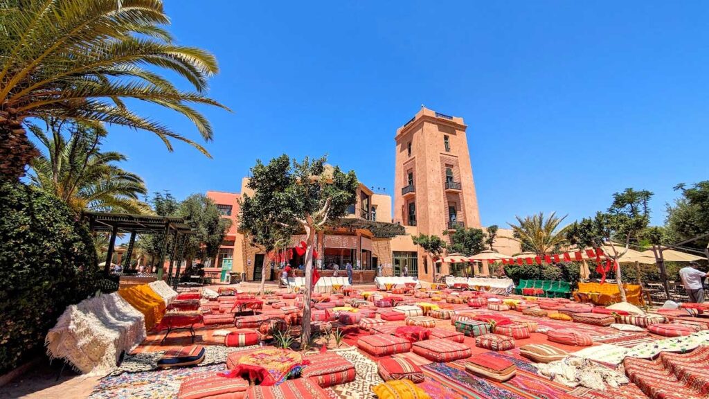 small red patterned pillows all over on the floor with tables. There are palm trees around the sitting area. There's a moroccan tower rising above into the sky