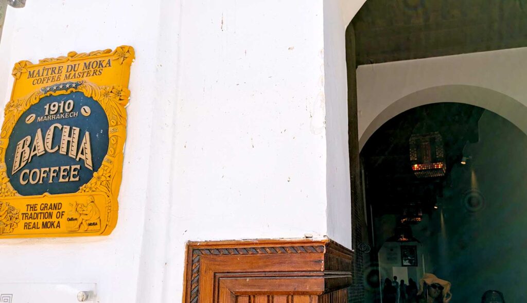 The outside entrance to bacha coffee. There's a yellow vintage sign on a white wall next to a rounded entryway that leads down a long hallway. It's dark in the hallway, but you can slightly make out people's backs as they wait in line, while others are walking out of the hallway, frustrated. 
The yellow vintage sign reads: Maitre du moka, coffee masters. In a black circle in the middle with yellow text: 1910 Marrakech are in between two coffee beans, Then Bacha Coffee. Underneath the black circle: The Grand tradition of real moka