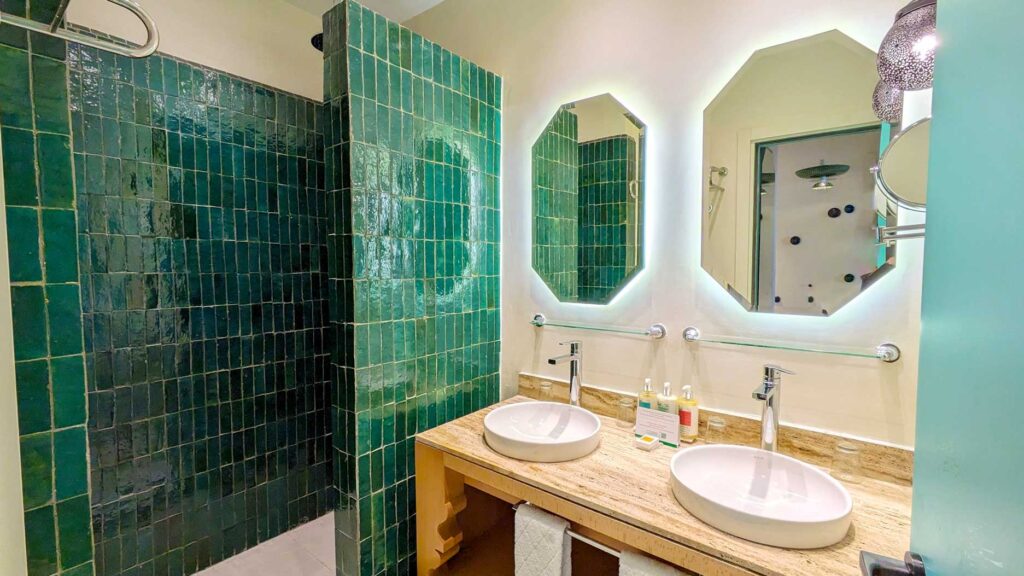 the bathroom at the club med la palmerie Marrakech in Morocco. the shower is an open shower, tiled with a rich evergreen glaze. There's a wood double sink vanity next to it with two hexagonal shape mirrors on the wall. There's three bottles in the middle.