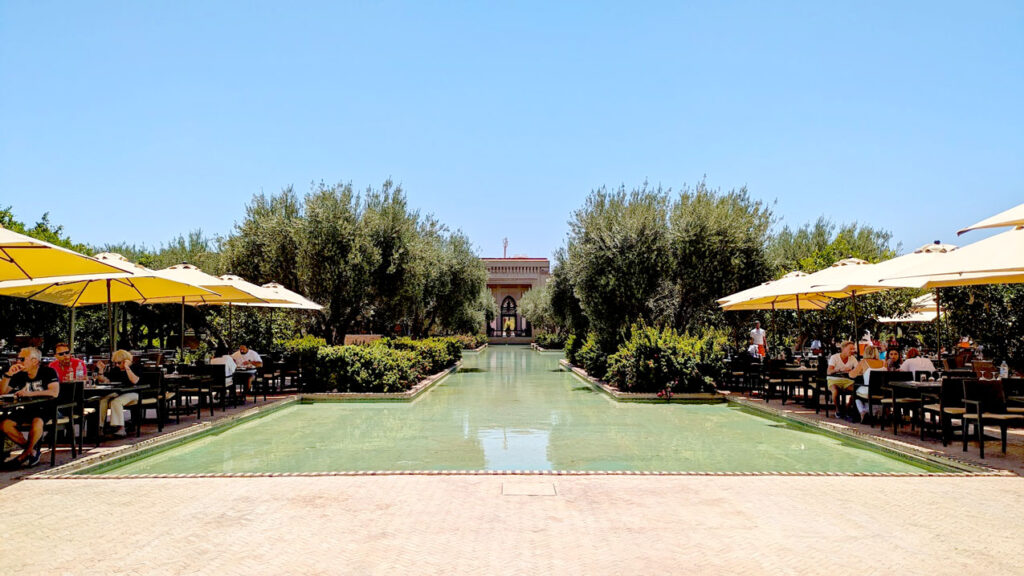 the view of the seating at the buffet at the club med in Marrakech. There are tables with umbrellas on either side of a shallow pool of water in the Marrakech gardens of bushes and olive trees. At the end you see the reception building with arched windows