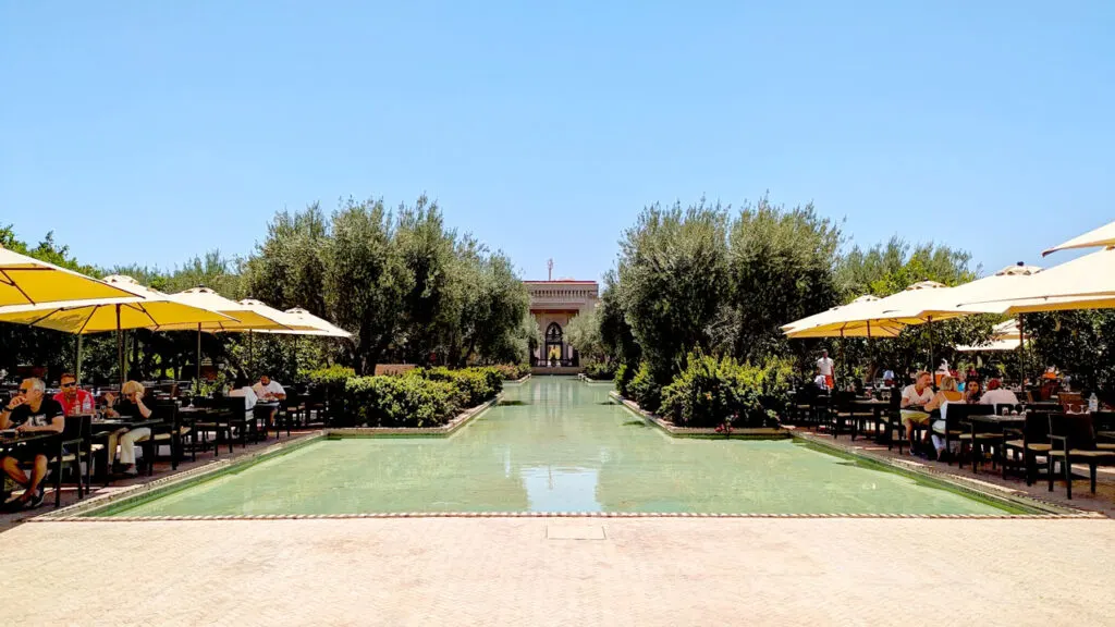 the view of the seating at the buffet at the club med in Marrakech. There are tables with umbrellas on either side of a shallow pool of water in the Marrakech gardens of bushes and olive trees. At the end you see the reception building with arched windows