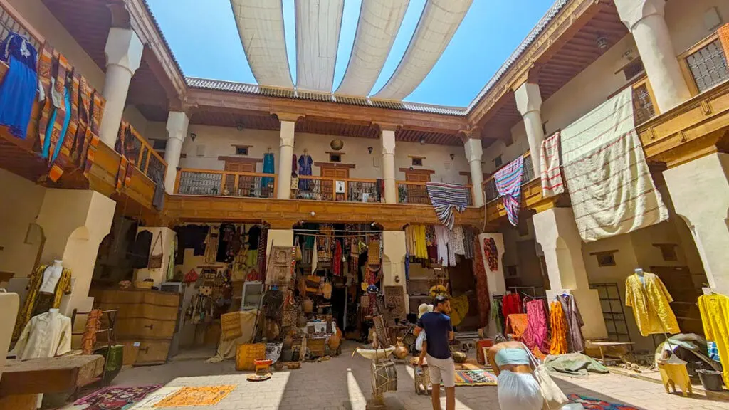 caravanserai example in the marrakech market. A courtyard off of the main market street. It has a two story building open to the center on every side. There's no roof, but there are four strips of fabric going across the top offering a bit of shade. there are a few items set up for sale