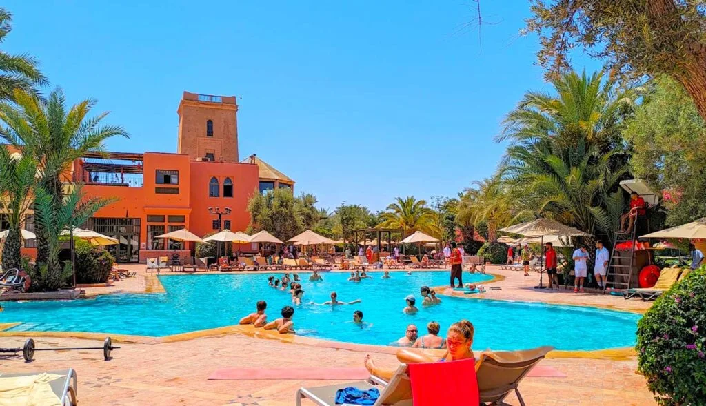 the pool at the club med marrakech in morocco. It's large, and there are quite a few people in the pool and sitting on pool lounge chairs around it. There are open umbrellas on the other side of the pool in front of a large moroccan style tower. There are short palm trees all around the pool