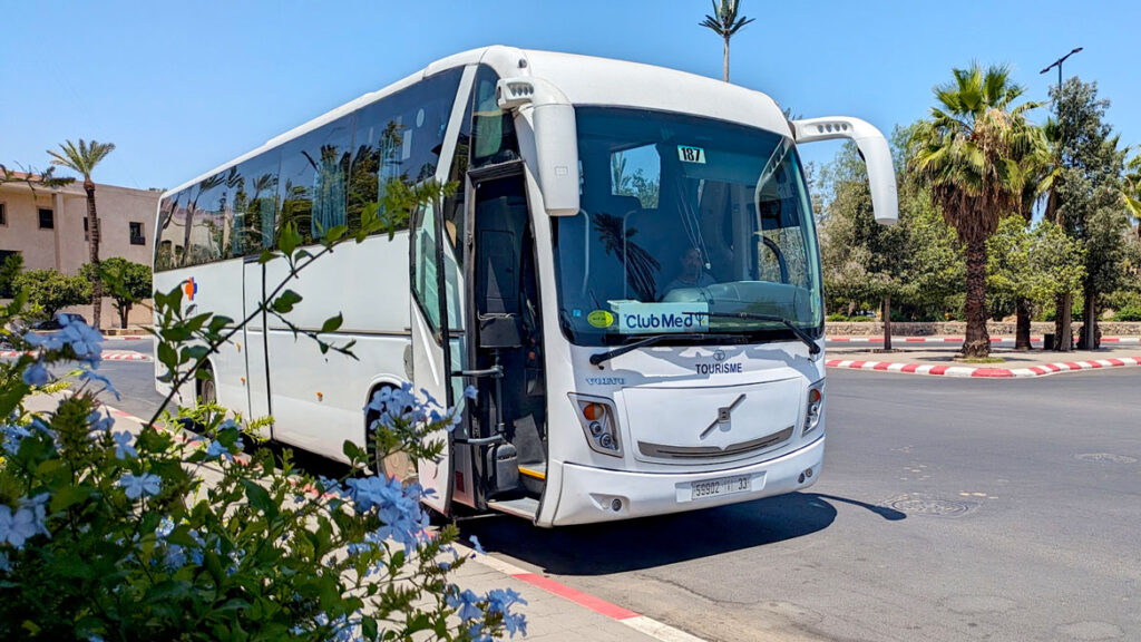 the club med shuttle bus is a large white greyhound bus, parked on the side of the street. There's a sign in the windshield that says club med with the logo. 