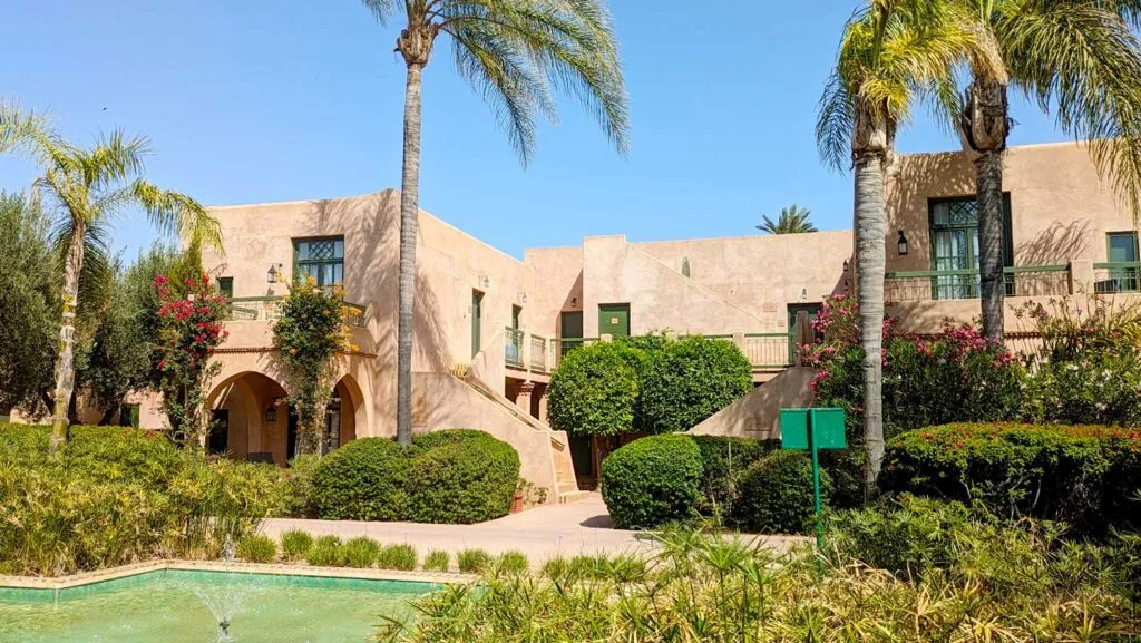 a building for the rooms at the club med la palmeraie marrakech morocco. A pale pink building, very square among palm trees and greenery. There's also a fountain in front of it. There isn't a cloud in the sky