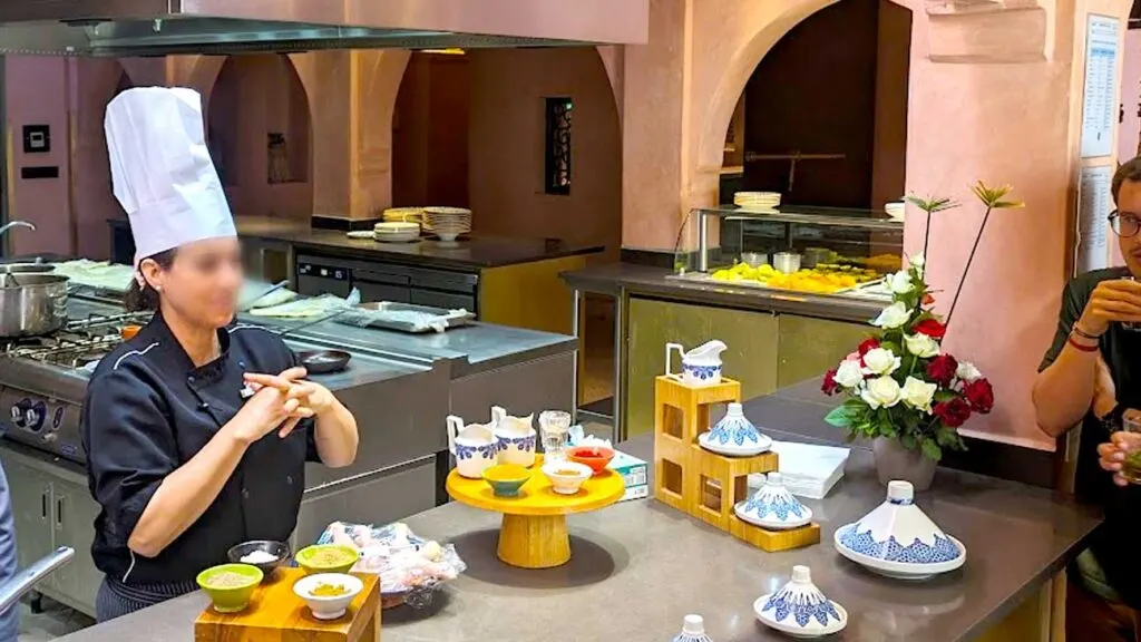 a woman wearing a chefs hat is standing behind an island speaking to a group of people. In front of her are several patterned tagines and pinch bowls filled with moroccan spices 