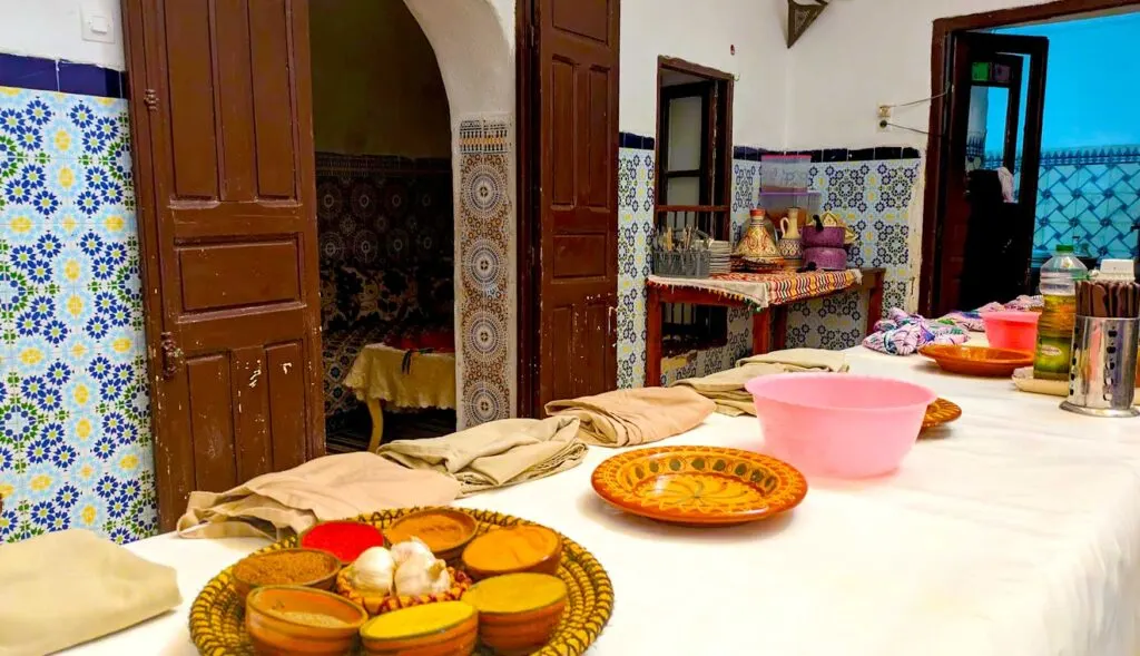 A long table set up in a traditional moroccan house all ready for the cooking class to begin. There are folded aprons set up on the left side of the table in spots for people to stand, underneath are cutting boards. In front of the aprons is a plate filled with pinch bowls for the different spices that will be used. Next to that sits an empty bowl, and finally there are jugs of oil at the end of the table