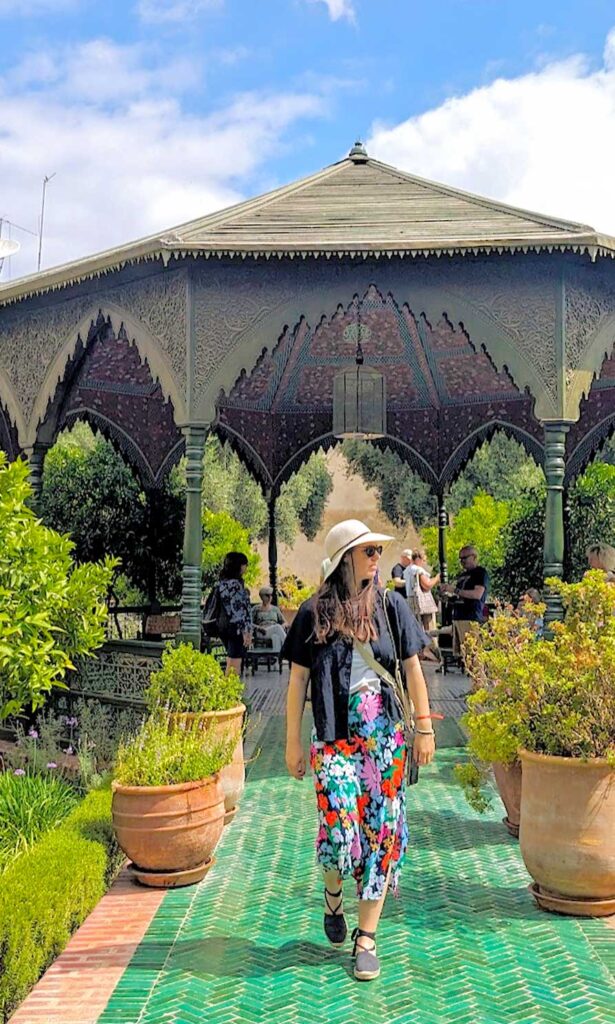 a woman walking tours the camera in the middle of a tiled walkway after walking out from a large gazebo. She's wearing espadrille sandals, a floral maxi skirt, a white tank top underneath a linen open buttoned down t shirt with a wide brim straw hag