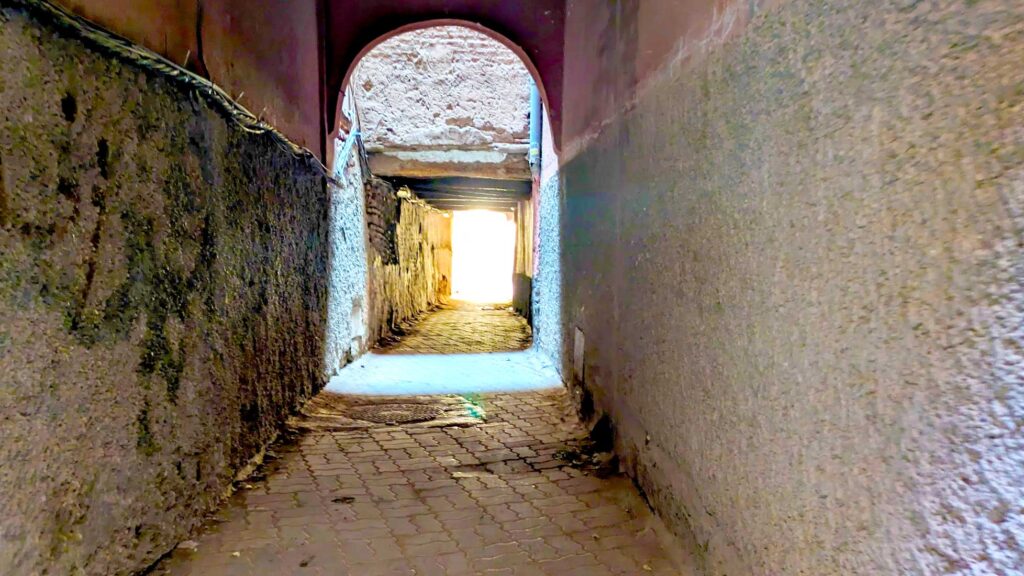 a creepy dark, ruined alley in the Marrakech markets. The ground is cement, but there are dirt piles near the walls. There's some graffiti on the wall. It's like a dark tunnel with some light at the end. You know you're still in Marrakech because the ceilings are rounded.