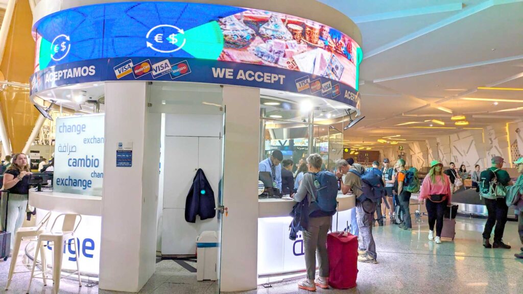 a circular currency exchange booth in the middle of the baggage claim at the airport. People are standing at the booths exchanging money. On the rim around the top it says they accept visa and mastercard, and $ and €
