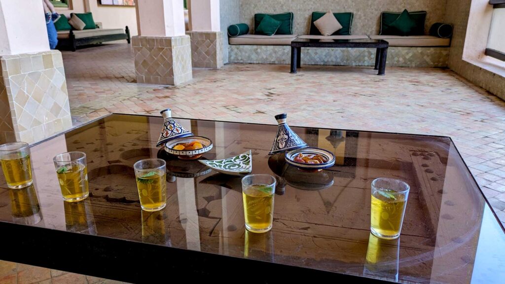 a glass top coffee table serves fives small glasses of moroccan mint tea, with fresh mint leaves floating inside. And two tagines filled with dates and dried apricots. The table takes up a large portion of the photo, but you can see the rough, unglazed, beige tile, and a couch and coffee table across the room