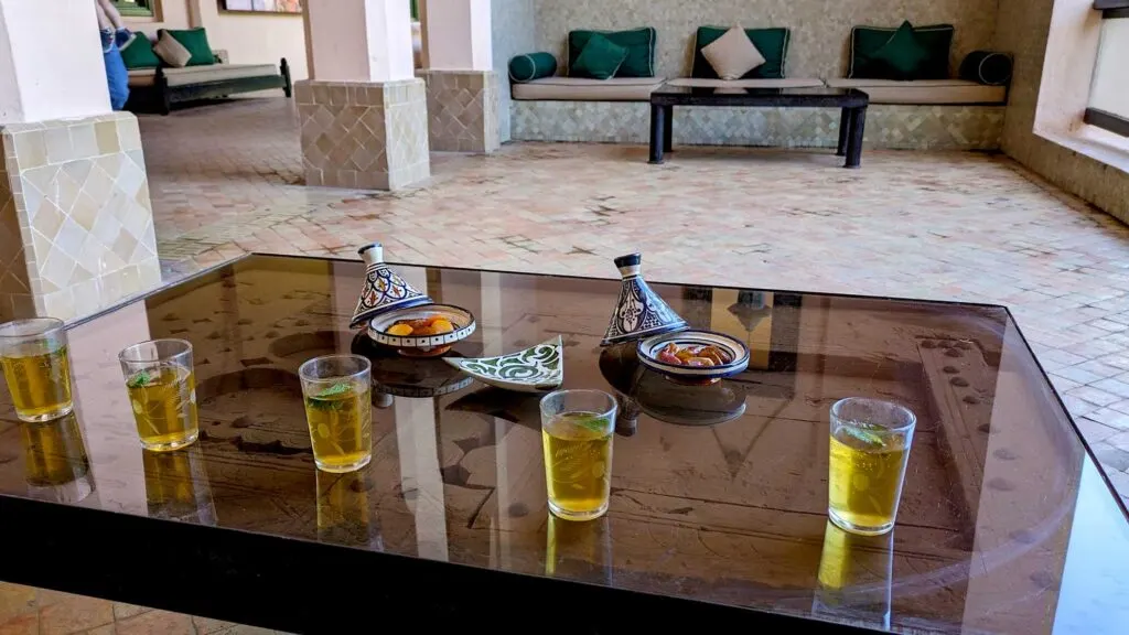 a glass top coffee table serves fives small glasses of moroccan mint tea, with fresh mint leaves floating inside. And two tagines filled with dates and dried apricots. The table takes up a large portion of the photo, but you can see the rough, unglazed, beige tile, and a couch and coffee table across the room