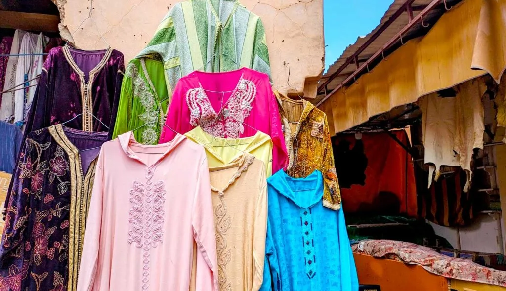 djeballas hanging on an outside wall of a market. Each one is a vibrant different colour with some decals on the front. There's a hot pink, bright blue, yellow, baby pink, and green