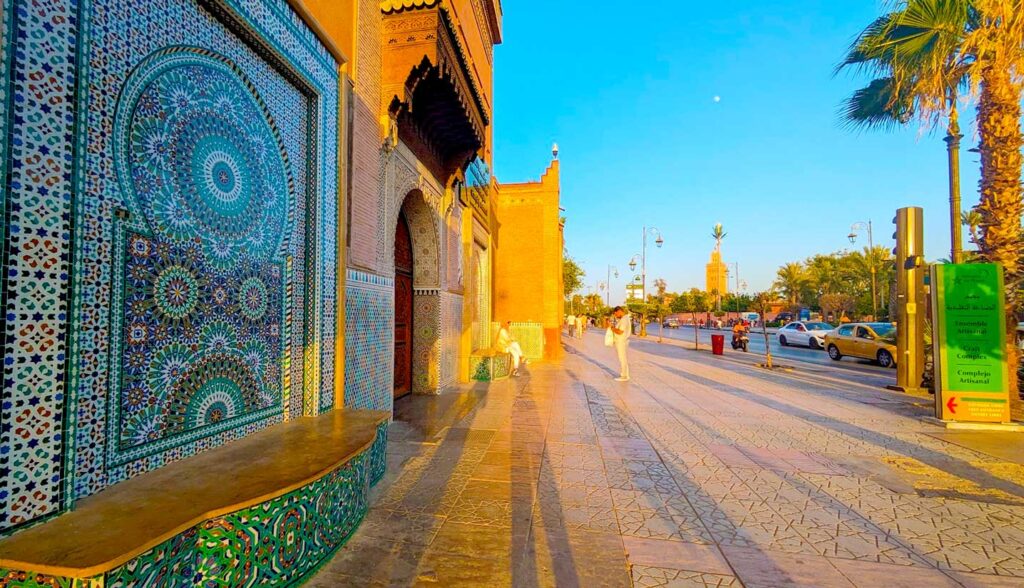 a view of the outside of the ensemble artisinal perfect for souvenir shopping in marrakech morocco. It's golden hour, so the side of the building is completely golden and people walking by and the trees create a long shadows on the sidewalk. 
There's a blue tiled mosaic with a bench on the left bottom of the photo. There's a bright green sidewalk sign opposite the door, it reads the Ensemble Artisinal. But the best part is that you can see the koutoubia mosque rising high above across the street in the distance