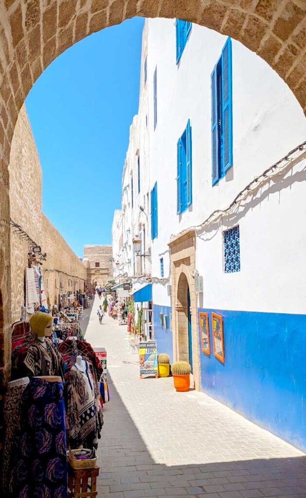 a view of the market shops in essaouira seen through an old grey brick arhcway. The archway matches the left wall of the alley and a tower at the end of the market. The right wall is completely different, it's white and blue painted stucco, from a newer era. Along the walls are shops that have placed their goods for sale outside along the walkway. People walk through the street looking at the shops. 