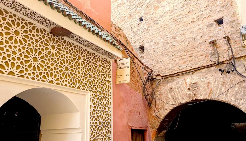 The outside of hammam mouassine. You wouldn't know but there's a small hanging sign off the side of the wall that has the name and Depuis since 1562. Underneath it says Pour Femmes, for women. The sign hangs on an old pink painted wall that leads into an ever older ruined brick wall connected to it with an archway leading to a dark tunnel. The entrance to the hammam is a wood carved mosaic that's been painted white