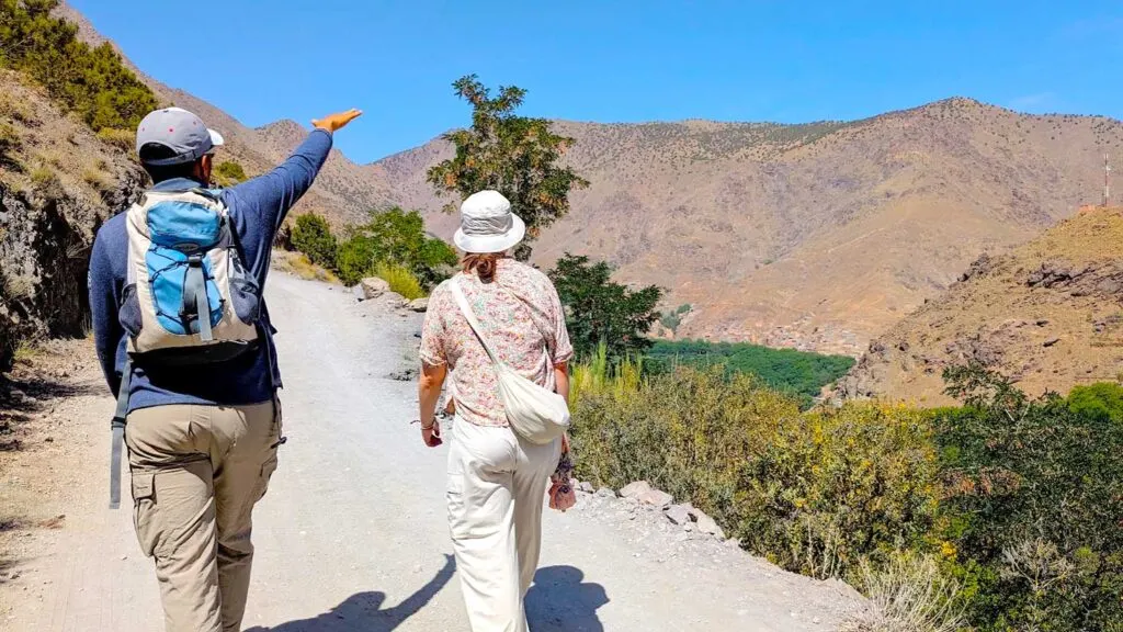 a man and a woman are photographed from behind walking on a gravel path with mountains surrounding them on every side. The woman is wearing beige palette with a floral blouse, a bucket hat, and full length pants. Even her purse matches the beige. The man is next to her, with his arm in the air, explaining something to the woman as they walk. He's also pretty covered up with long sleeves and pants.