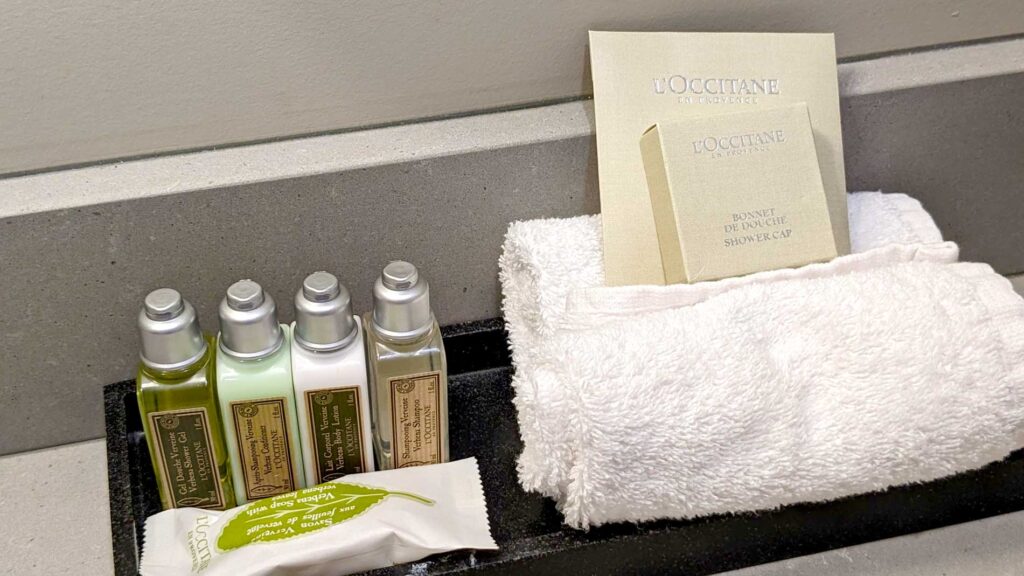 a closeup of bathroom hotel essentials. these are from the brand Occitaine, featuring a shampoo, conditioner, body lotion, and shower gel, as well as a sealed bar of soap in packaging. There are two towels rolled up with a packed shower cap resting on top