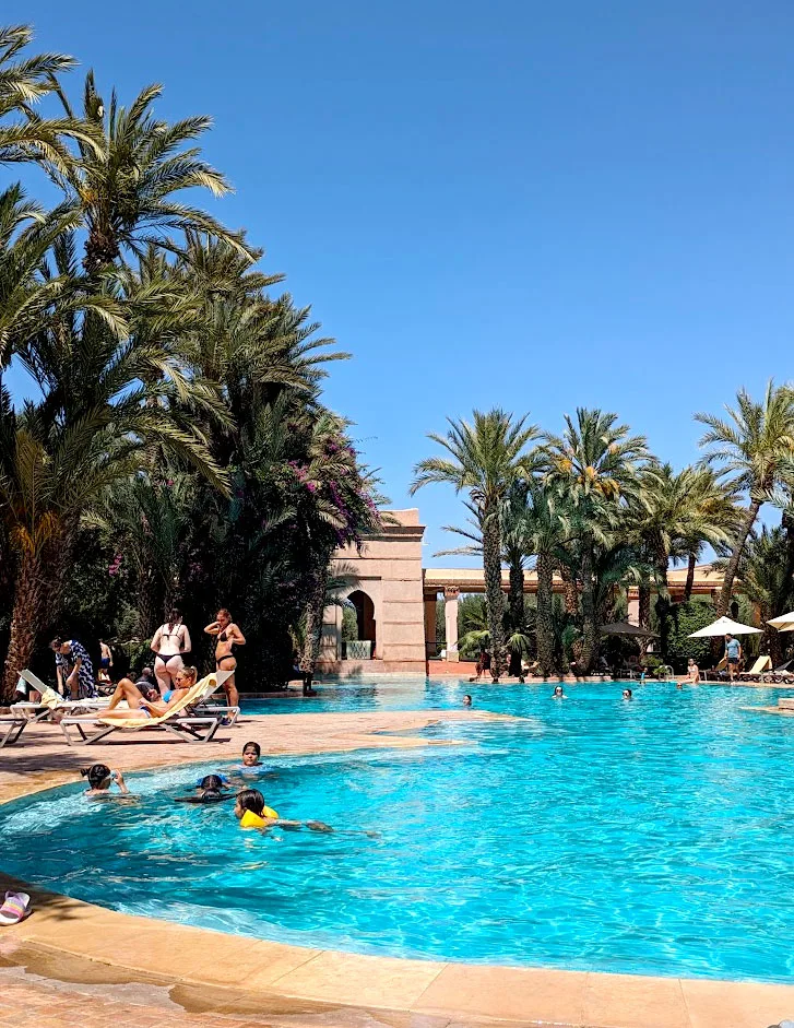 a vibrant blue pool that's huge. It leads all the way to a moroccan archway. Palm trees and lounge chairs line the outside of the pool. There are a few groups in the pool, other people are lounging outside on the chairs