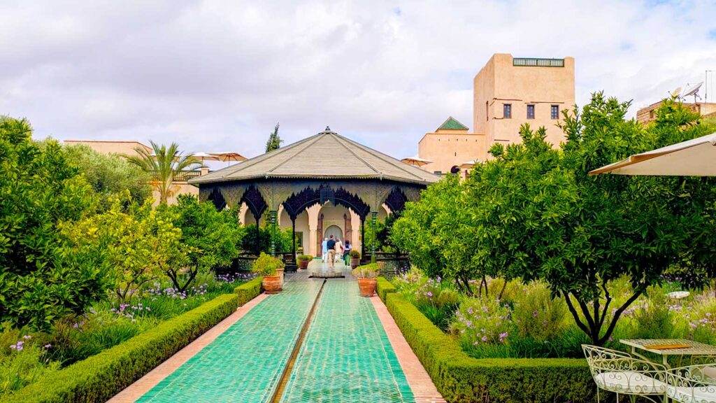 the view of the secret garden with all the highlights. A green tiled path leads you off into a covered gazebo in the centre of garden. Of to the right, there are romantic white metal garden chairs with a table and umbrella. On either side of the pathway are small trees, probably fruit trees that are out of season and flowered bushes below.  Behind the veranda is a tower, that looks like it has a rooftop with a barrier. 