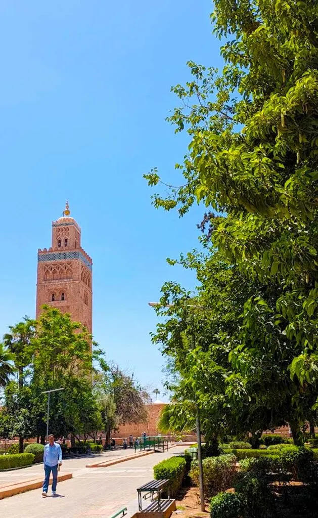 the view of the koutoubia tower rising in the sky from the koutoubia gardens. On the right side, is a tall tree that cuts off by the top of the image. There's a pathway leading to the koutoubia grounds, a solo man is walking in the middle.  But you can see more people walking closer to the koutoubia