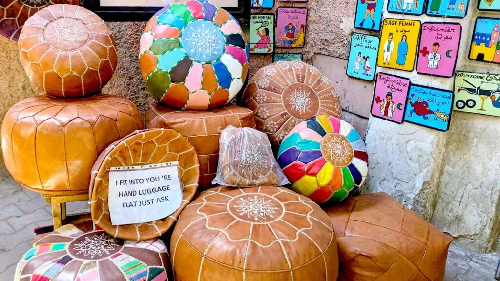 leather poofs sit on the floor outside a shop in a marrakech market. The poofs all have the same design, and are a creamy brown colour with a creamy white stitching. One of the poofs is completely flat, with no stuffing, there's a printed paper on it that says: I fit in you hand luggage flat, just ask