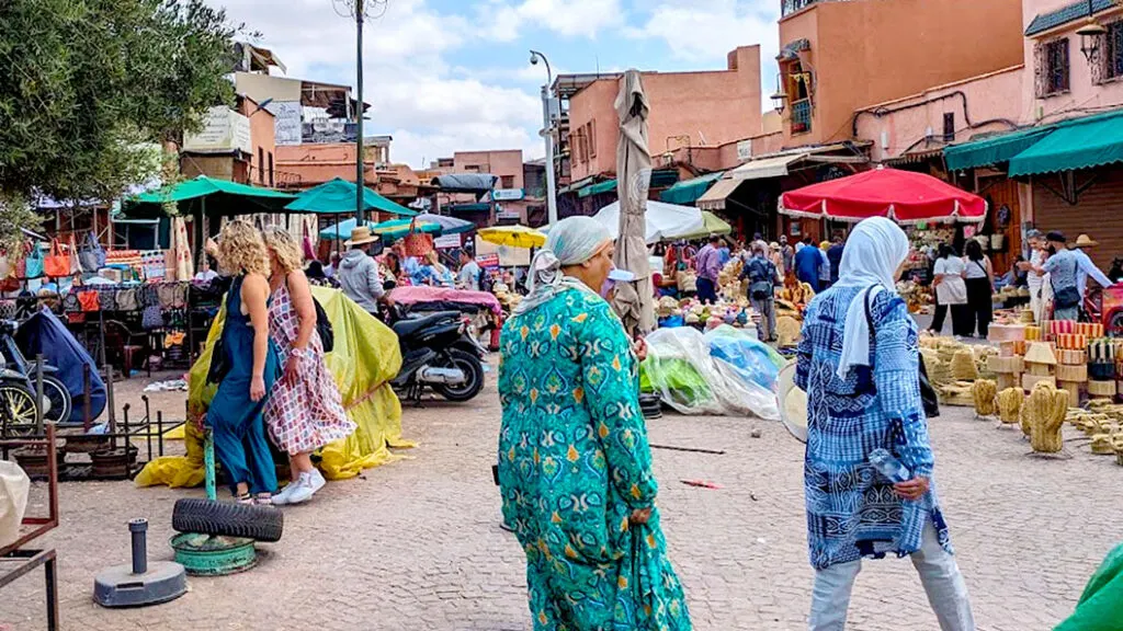 a very marrakech market scene. It's a bustling square with souvenirs on the ground for sale, umbreallas are set up throughout the square for shade, red square buildings like the right side of the square, and people are walking in every direction.

But it't the two women walking by in front of the camera that mainly catch your eye. An older woman, in her 50s, is wearing a long sleeve, long dress with a green pattern. It's long because she's holding up the bottom of it in her hands. Her head is covered with a wrapped scarf. 

In front of her is another woman, but you can't see her face, she has a scarf covering her head and neck. She's wearing a long shirt that goes down to her knees, with long sleeves with a blue and brown pattern, She's always wearing white pants. 