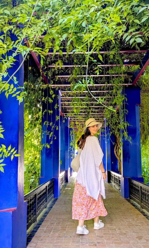 A woman walking forward but looking back under a completely covered pathway by leaves. There are vibrant blue beams holding up the leaf roof. The woman is wearing a straw sunhat, a white loose kimono, and a long pink and orange maxi dress that just barely shows her ankles before you see her white sneakers