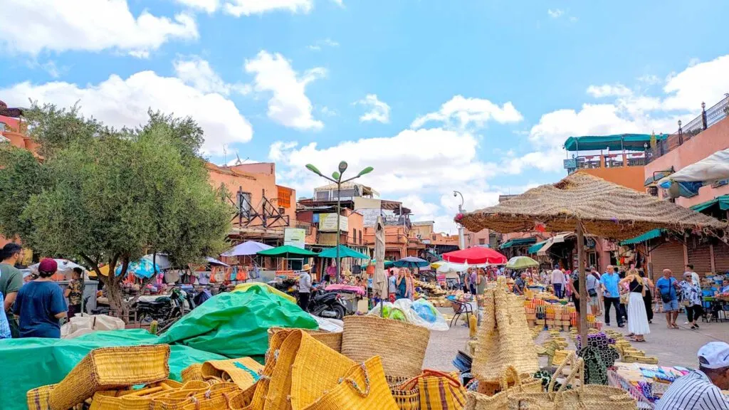 the bustling square in the middle of the mazes of the marrakech market. There are straw bags for sale in the bottom of the image. Beyond that everything melds together: people walking, shopping, and selling goods. There are umbrellas set up for shade. Buildings, with rooftop terraces, along the edges and in the background. There's a lonely tree standing out in the centre of it all.