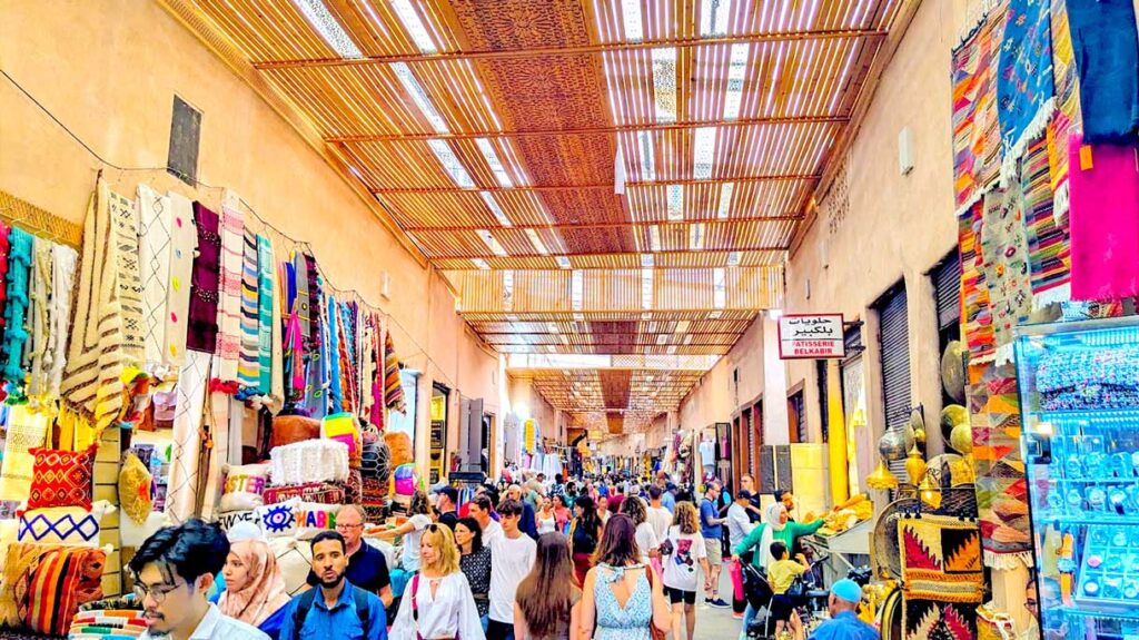 the market in Marrakech. It's a covered thatched roof letting in some sunlight. Vendors are selling goods on either side of the alley, but it is packed with people the entire way down