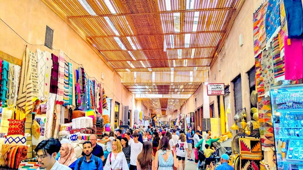 the market in Marrakech. It's a covered thatched roof letting in some sunlight. Vendors are selling goods on either side of the alley, but it is packed with people the entire way down